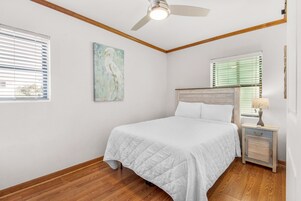 Un dormitorio con cama, ventilador de techo, mesita de noche con lámpara y ventana con persianas.