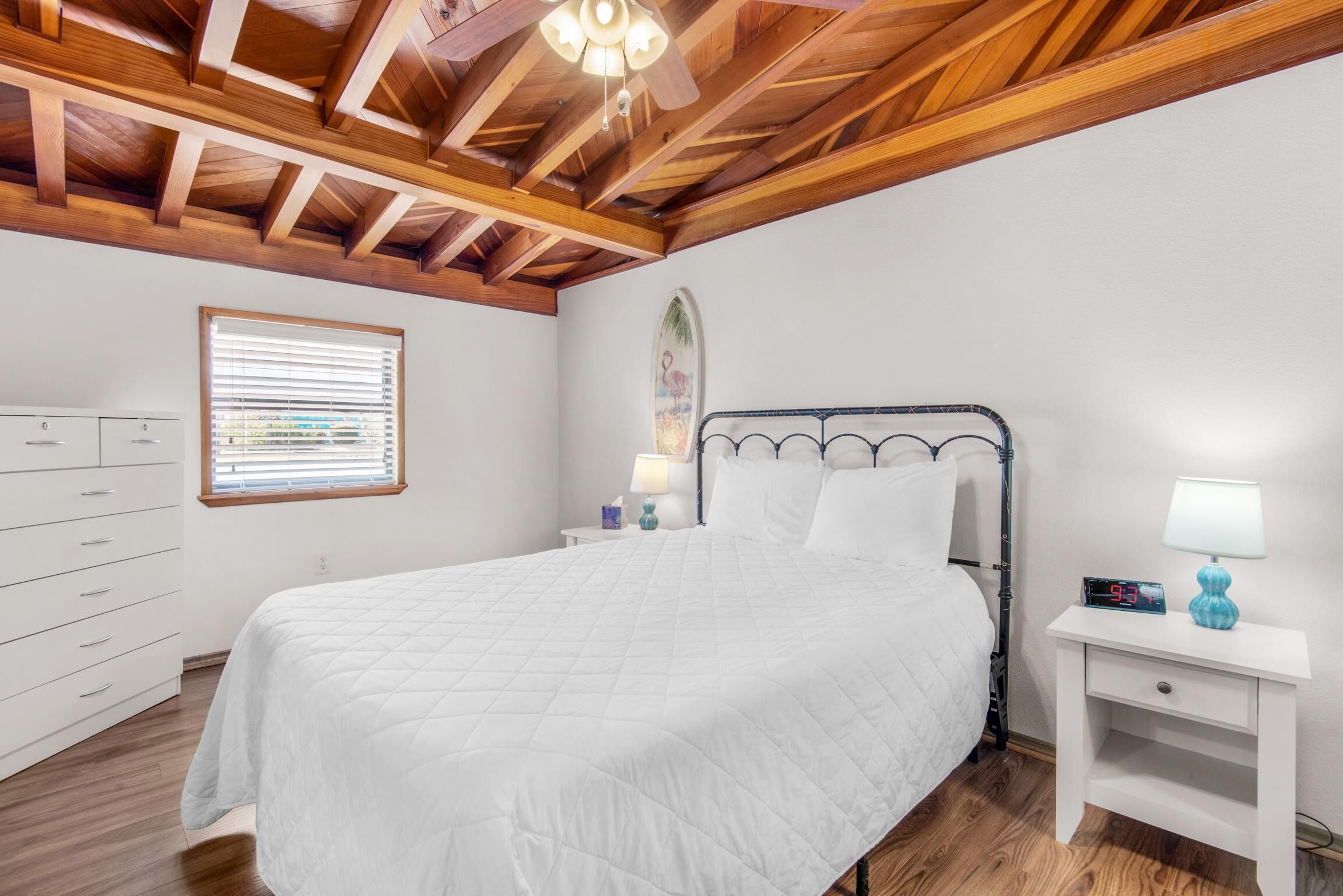 A bedroom with a bed, a nightstand, a dresser, a window with blinds, and a wooden ceiling.