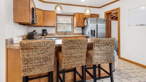 A kitchen with wooden cabinets, a stainless steel refrigerator, a coffee maker, and three wicker bar stools. - The Sugar Shack (Pensacola Beach)