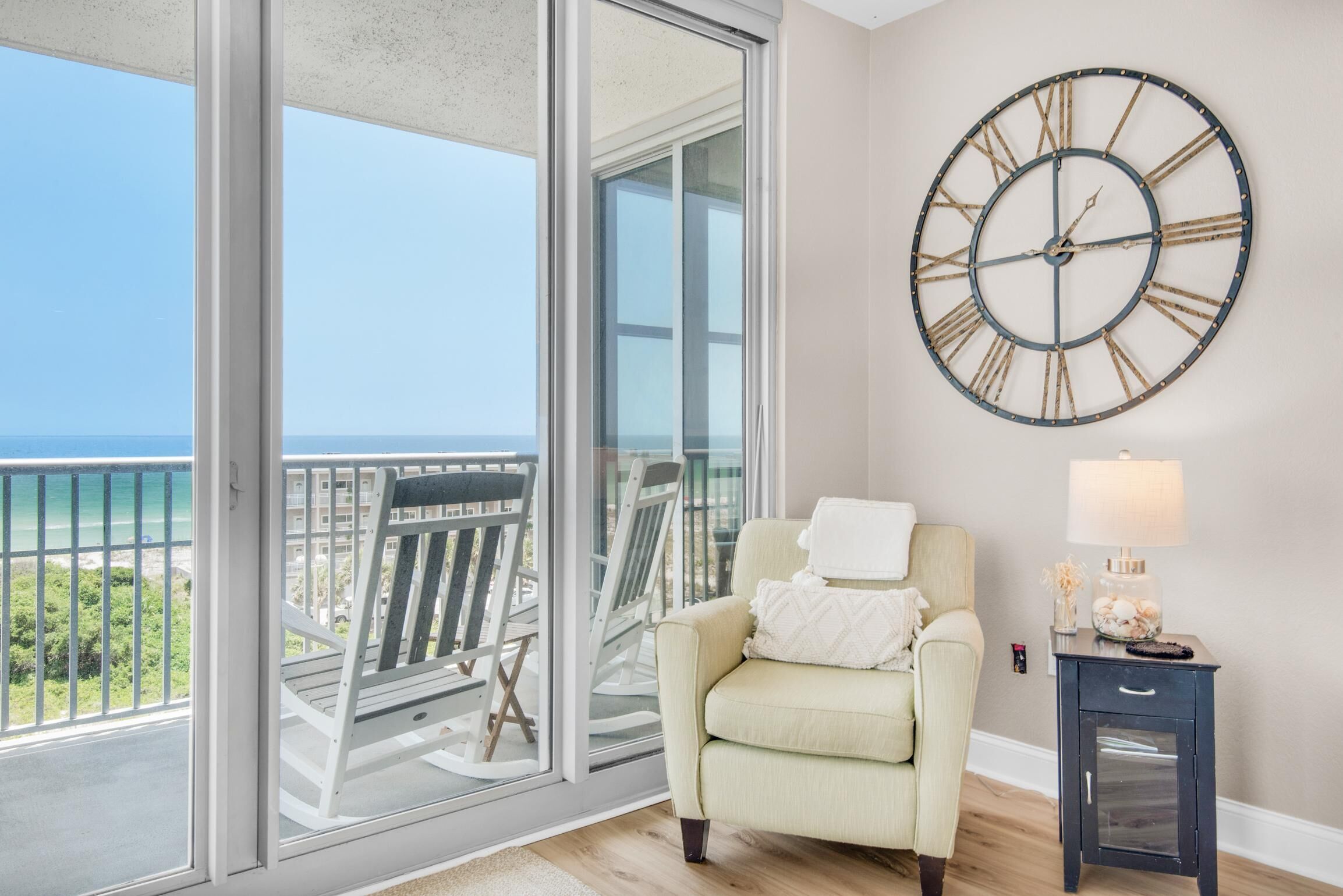 A room with a large window, a chair, a small table with a lamp, and a wall clock.
