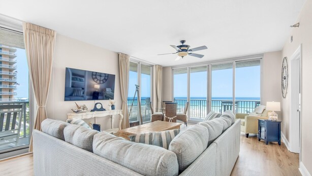 A spacious living room with a large gray sofa, a wooden coffee table, and a television mounted on the wall. There is a balcony with a view of the ocean.