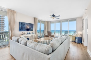 Sala de estar espaçosa com um sofá cinzento grande, uma mesa de centro de madeira e uma televisão fixada na parede. Há uma varanda com vista para o mar.