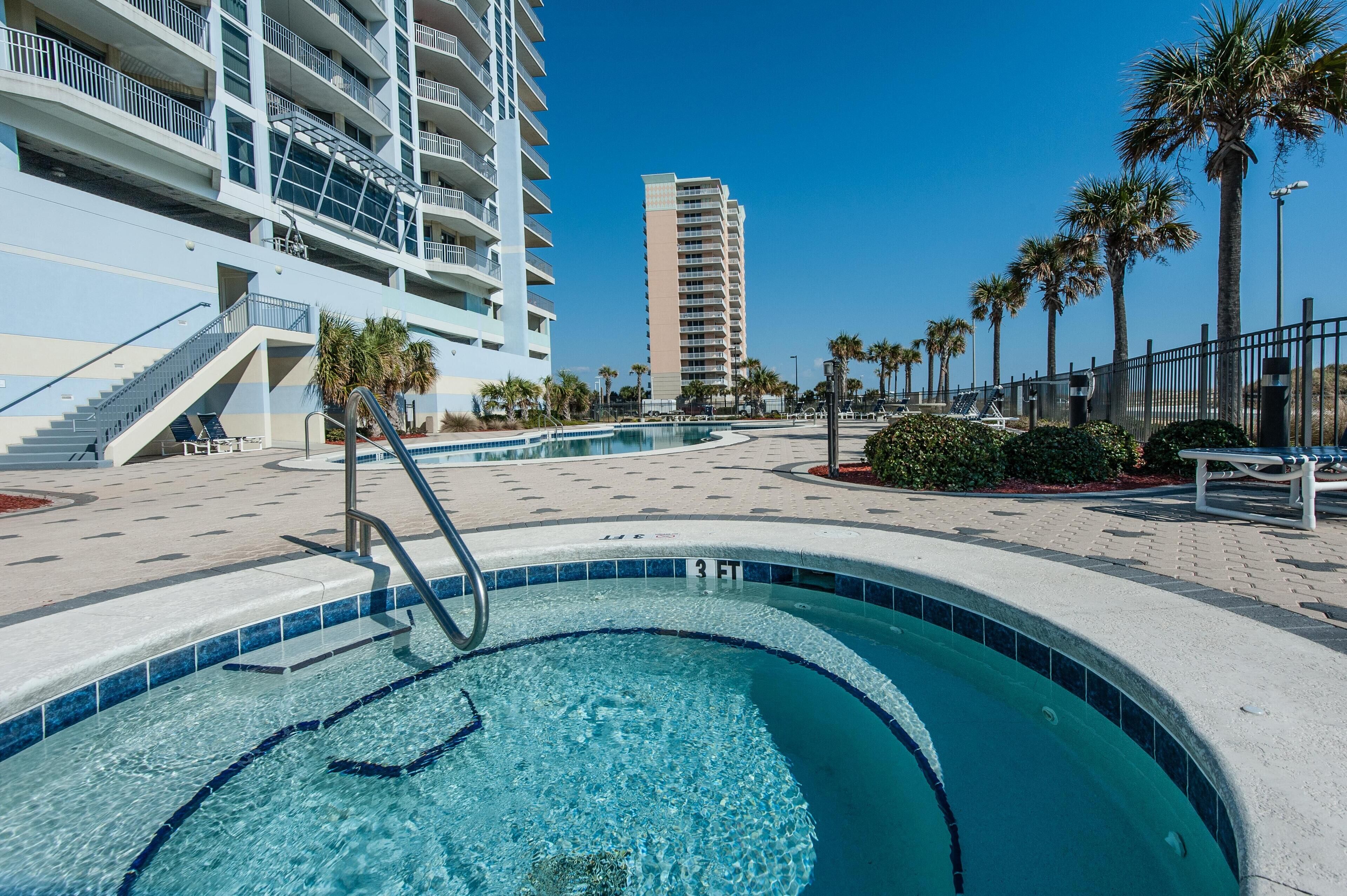 A swimming pool area with a clear blue pool, steps leading into the pool, a palm tree-lined walkway, and a high-rise building in the background.