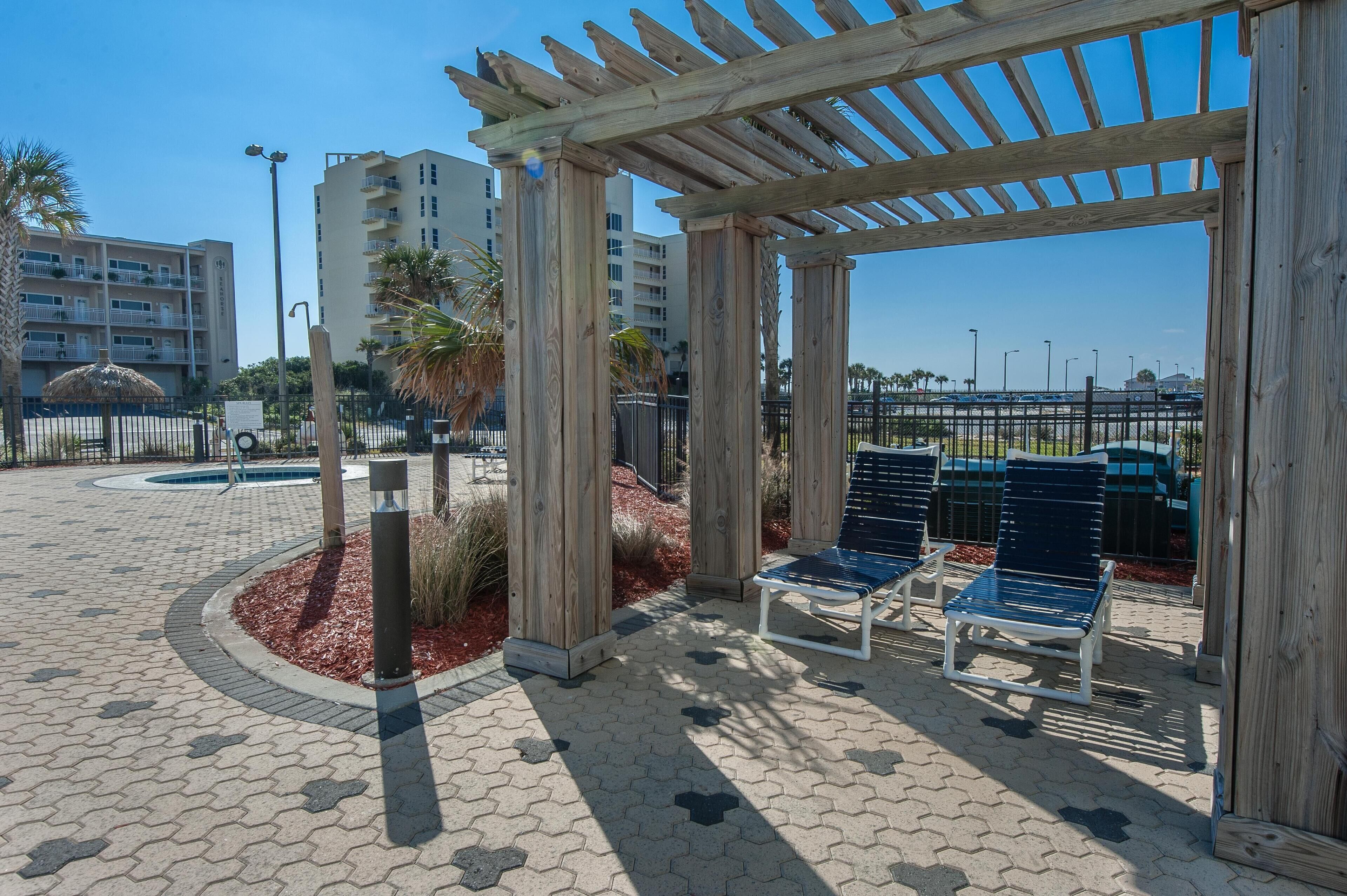 A covered outdoor seating area with wooden pillars, a paved walkway, and a view of a parking lot and buildings in the background.