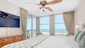 A bedroom with a large bed, a ceiling fan, a TV, and a view of the beach.