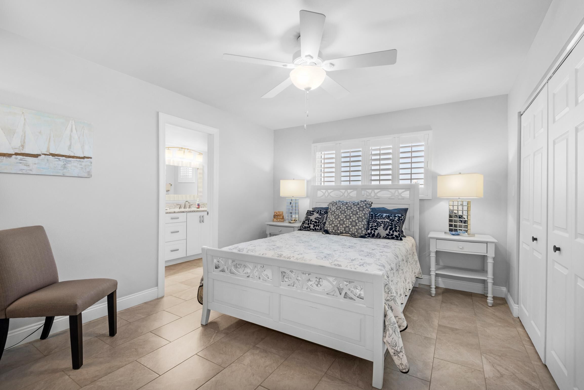 A bedroom with a white bed, two bedside tables with lamps, a ceiling fan, a chair, and a door.