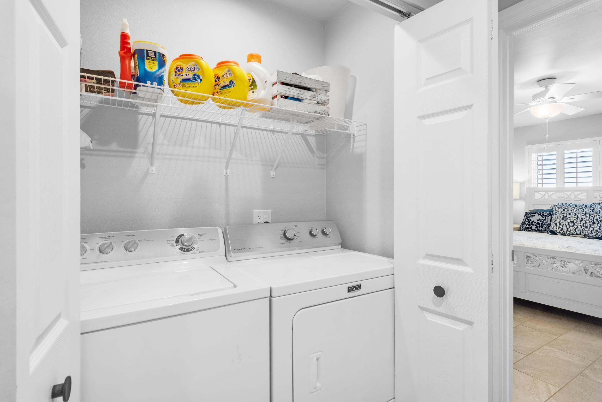 Un espace buanderie équipé d’un lave-linge et d’un sèche-linge à chargement frontal, d’une étagère avec des bouteilles de lessive, et une chambre adjacente avec un lit recouvert d’une literie à motifs.