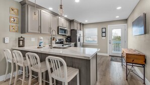 A modern kitchen with a large island, white bar stools, and a dining table with chairs. - Deep Water Cove 103 - OLD (Pensacola Beach)
