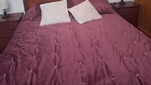 Un lit avec une tête de lit en bois, deux coussins décoratifs, une couvre-lit rouge matelassée, une table de chevet avec une lampe et une armoire.