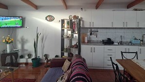 Un salon avec un canapé, un téléviseur diffusant un match de sport, un coin repas avec une table et des chaises, et une cuisine avec des meubles blancs et un dosseret carrelé.