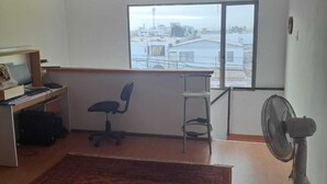 Une pièce comprenant un bureau, un fauteuil de bureau et un ventilateur sur pied. Une grande fenêtre offre une vue sur des bâtiments et une étendue d’eau.