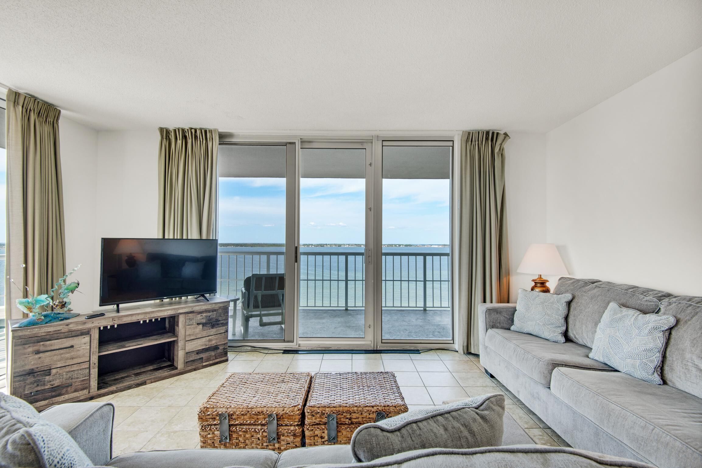 Un salon doté d’un téléviseur à écran plat, d’un canapé gris, de poufs en osier et d’un balcon offrant une vue sur la mer.