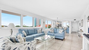 A spacious living area with two blue sofas, a glass coffee table, and a view of the cityscape through large windows. - Santa Rosa Dunes 1021 (Pensacola Beach)