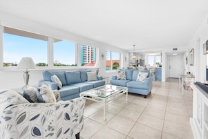 A spacious living area with two blue sofas, a glass coffee table, and a view of the cityscape through large windows.