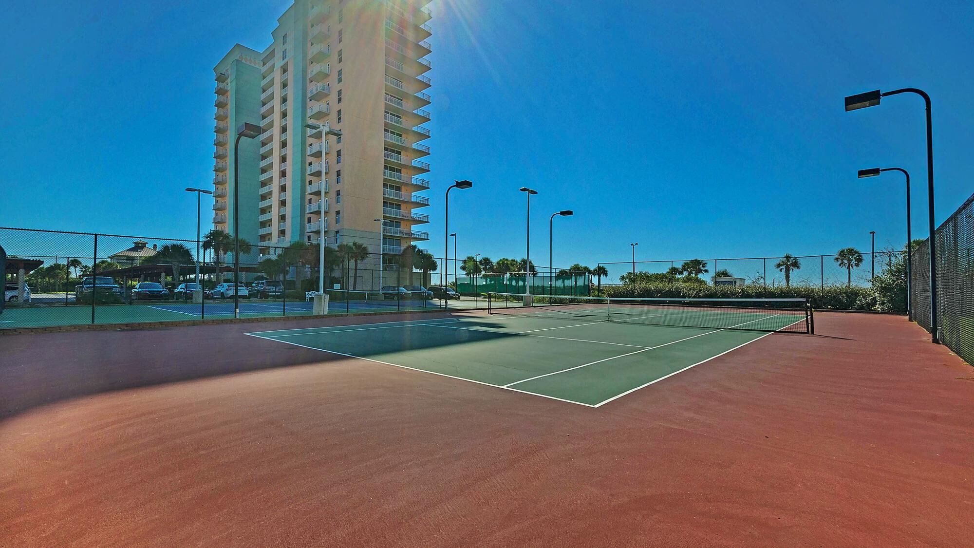 A tennis court with a net, surrounded by a fence, and a tall building in the background.