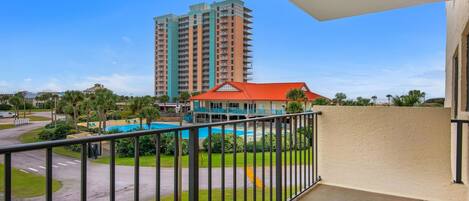 A balcony with a view of a swimming pool, a tall residential building, and a red-roofed building.