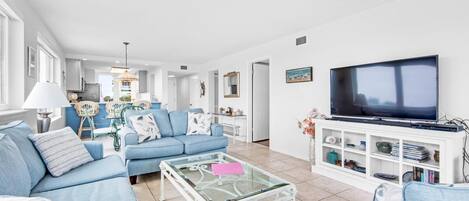 Una sala de estar moderna con un sofá azul, una mesa de centro de vidrio, un televisor sobre un mueble blanco y una cocina al fondo.