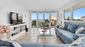 A modern living room with a glass coffee table, a sofa, a TV, and a view of a balcony with a pool. - Santa Rosa Dunes 1021 (Pensacola Beach)