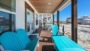 Un espacio al aire libre techado con suelo de madera, sillas y mesas azules, con vistas a una playa y edificios.