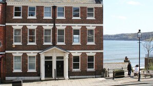 A two-story brick building with a white trim facade, a central entrance with a pediment, and a view of the sea in the background. - Waimea Apartment 4 | Best Sea View, Beach Front, Parking, Scarborough (North Yorkshire)