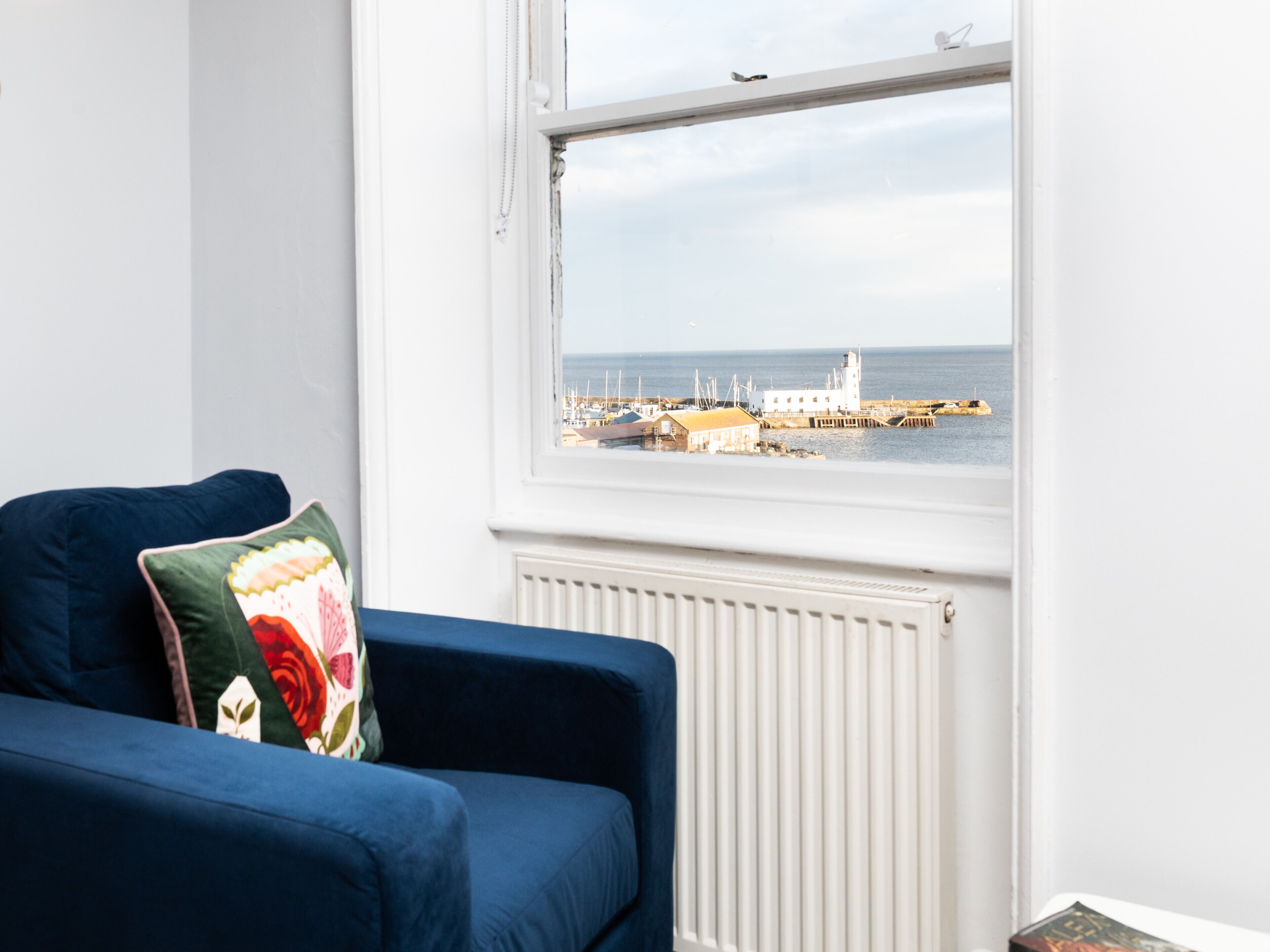 A room with a blue sofa and a window overlooking a harbor with boats and a white building.