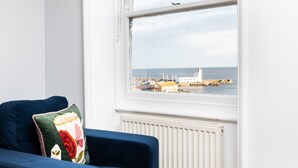 A room with a blue sofa and a window overlooking a harbor with boats and a white building. - Waimea Apartment 4 | Best Sea View, Beach Front, Parking, Scarborough (North Yorkshire)