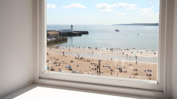 A beach view through a window, including a pier and a lighthouse. - Waimea Apartment 4 | Best Sea View, Beach Front, Parking, Scarborough (North Yorkshire)