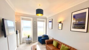 Una sala de estar con un sofá, una mesa de centro y un televisor. Se ven dos ventanas con vista a la playa.