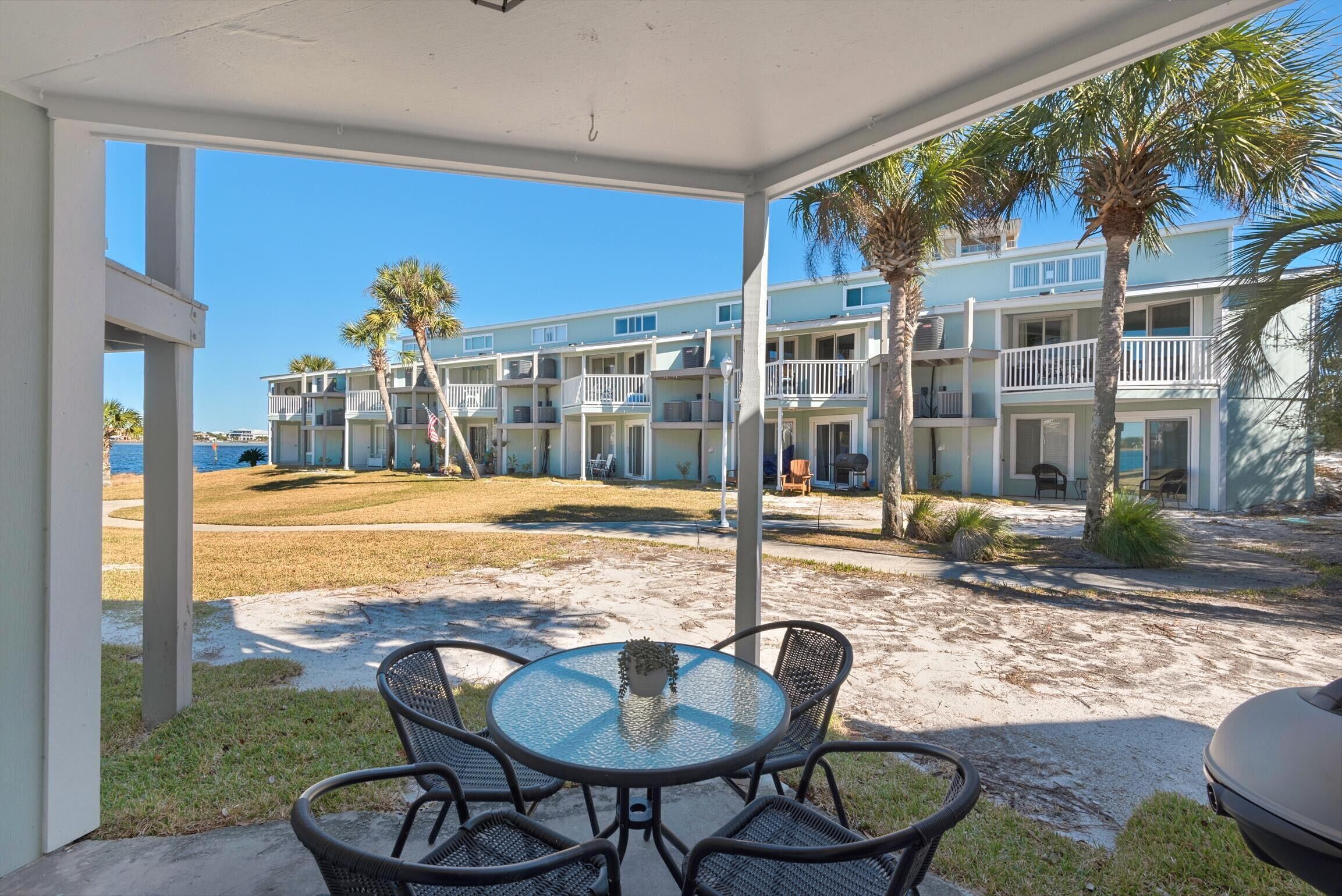 Un espace extérieur aménagé, abrité et doté d’une table et de chaises, offrant une vue sur un bâtiment situé en bord de mer, agrémenté de plusieurs balcons et de palmiers.