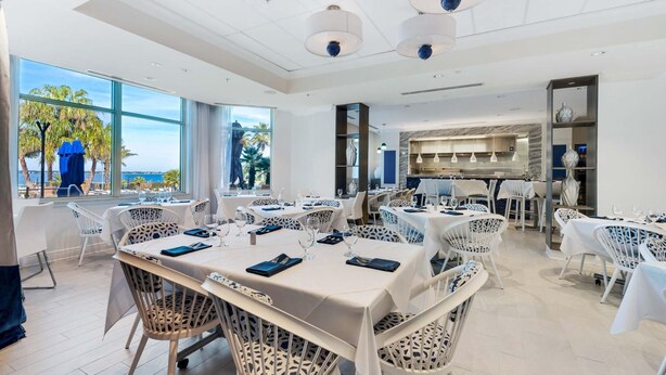 A spacious dining area with multiple tables and chairs, large windows offering a view of palm trees and the sea, and modern pendant lighting.
