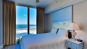 A bedroom with a large bed, a nightstand, a lamp, and a painting of a beach scene.