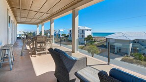A covered outdoor area with wicker furniture, a table, and a view of the beach and ocean. - Emerald Oasis (Pensacola Beach)