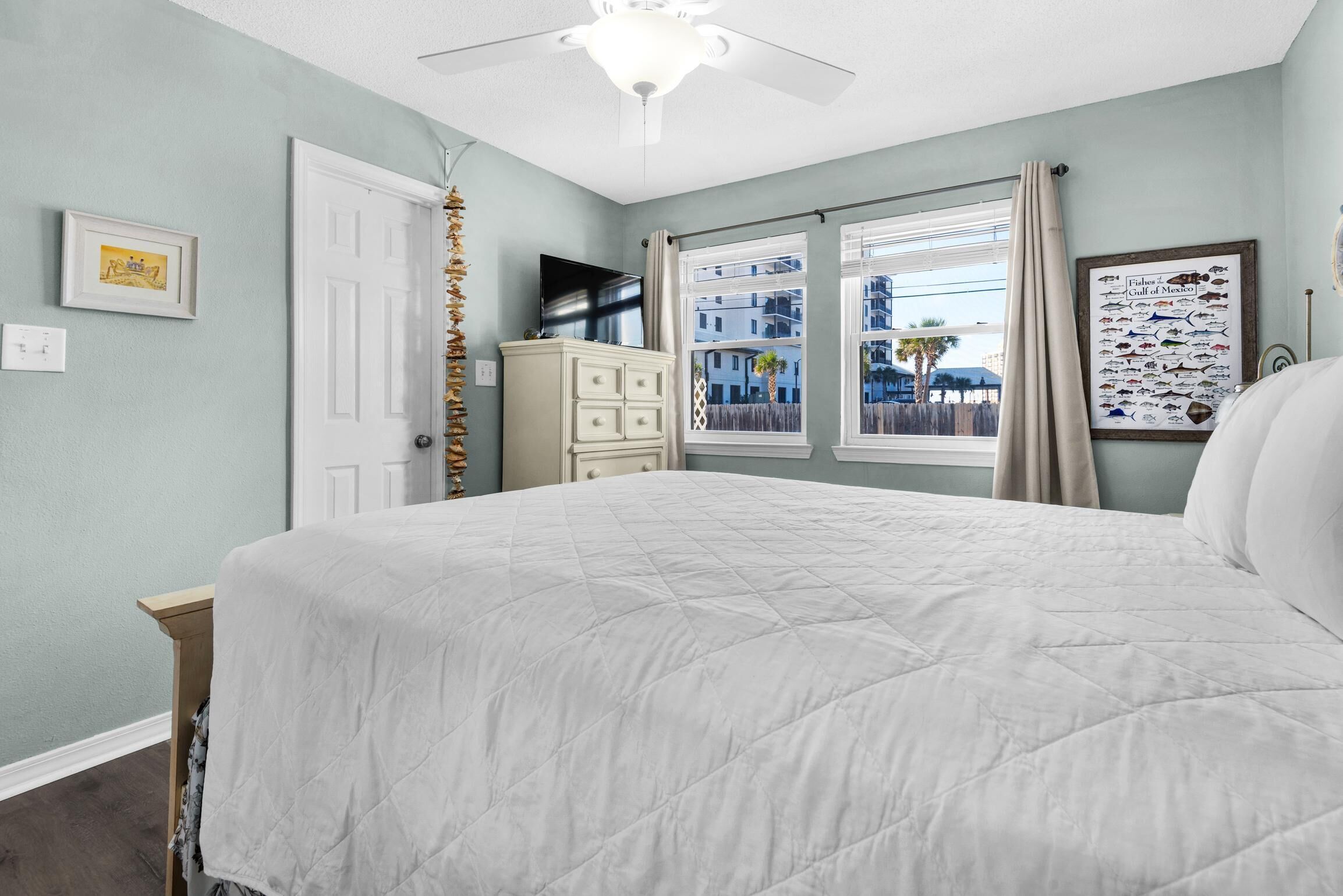 A bedroom with a large bed, a ceiling fan, a dresser, a TV, and a window with a view of buildings and palm trees.
