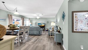 A spacious living room with a grey sofa, a dining table with chairs, a TV, and a wall-mounted shelf. - Beachview 101- Beaches and Moonbeams (Navarre)