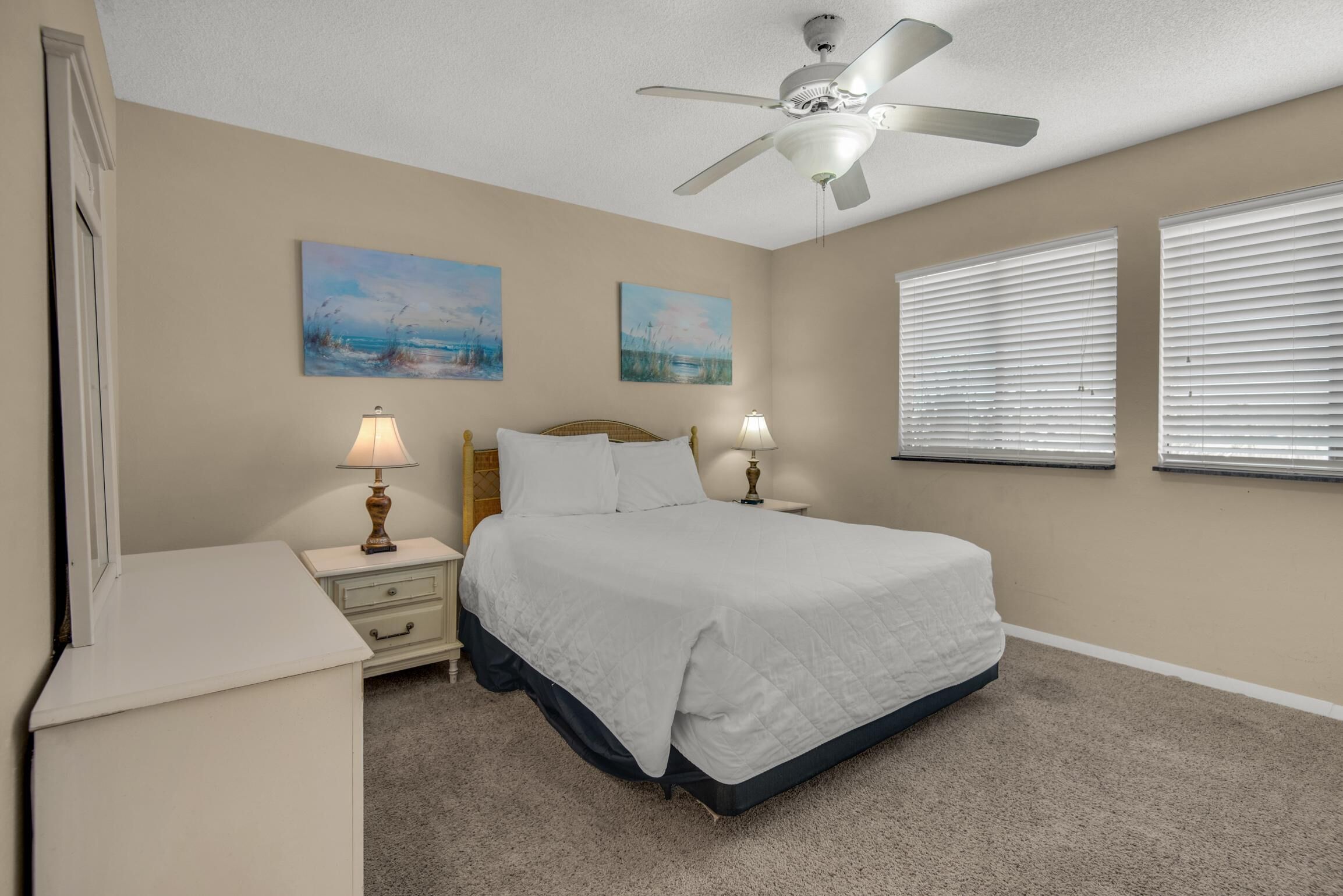 A bedroom with a bed, nightstands, lamps, ceiling fan, artwork on the wall, and windows with blinds.