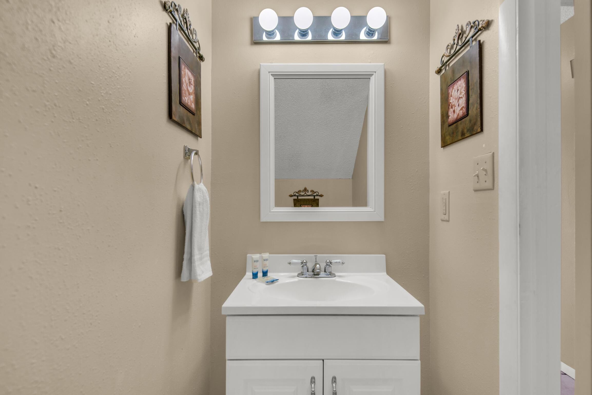 A bathroom with a white sink, vanity, and a mirror with lights. There are framed pictures on the walls and a towel hanging on the left.