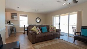 A living room with a brown sofa, a coffee table, and a patterned rug. There is a kitchen area with cabinets and a dining table. - Sunset Harbor Villas 431 (Navarre)