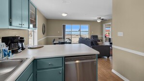 A kitchen with a dishwasher, a dining area with a table and chairs, and a view of the beach through large windows. - Sunset Harbor Villas 431 (Navarre)