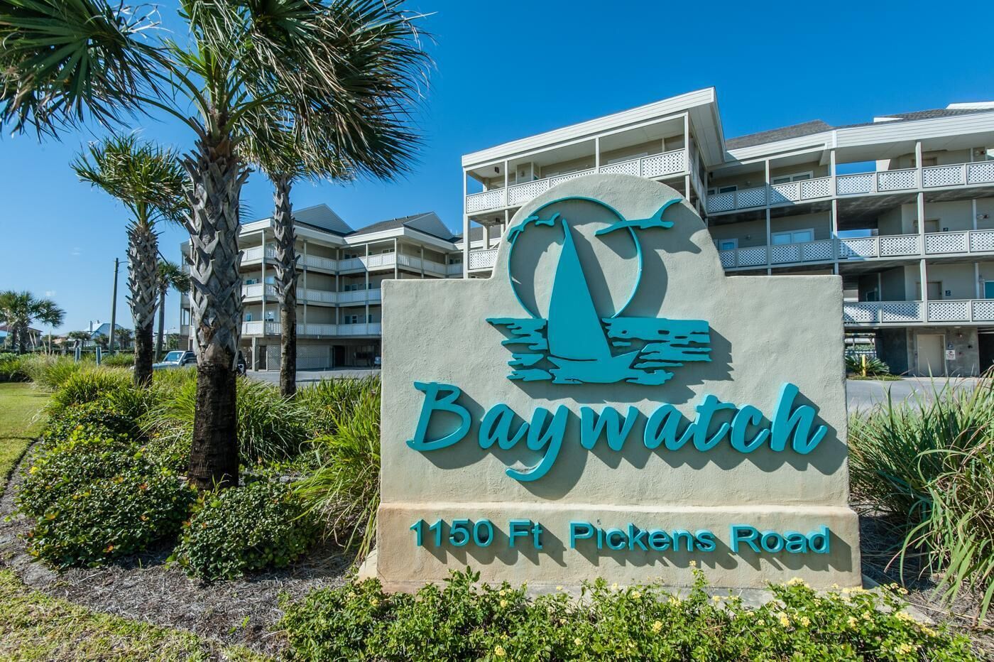 A sign for Baywatch at 1150 Ft Pickens Road with a building in the background.