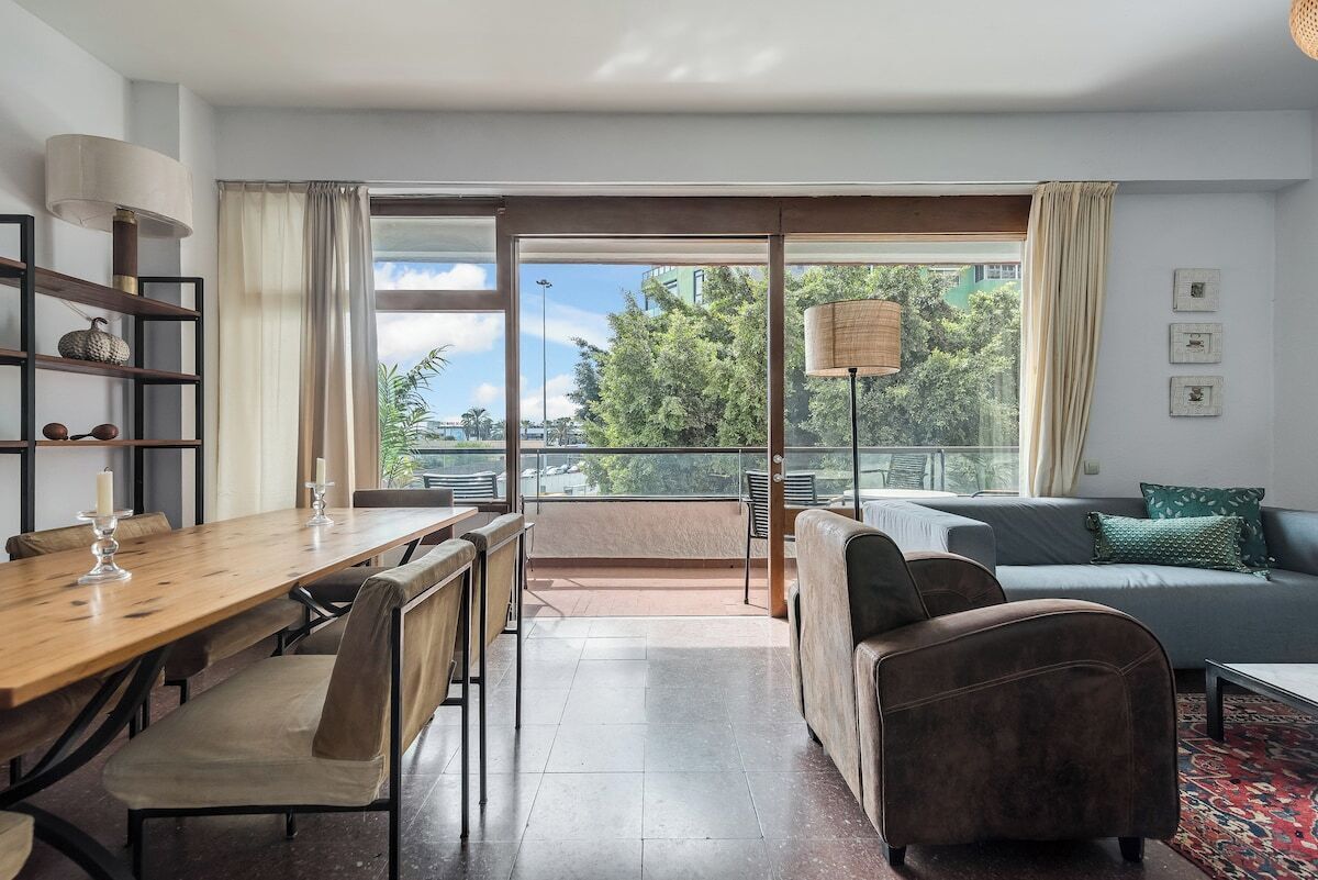 A modern living room with a wooden dining table, cushioned chairs, a sofa, and a balcony with a view of greenery.