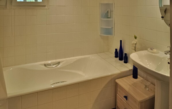 A bathroom with a bathtub, a sink, and a mirror. There are three bottles on the floor and a vase on the wall. - Lovely home in Lacoste (Lacoste)