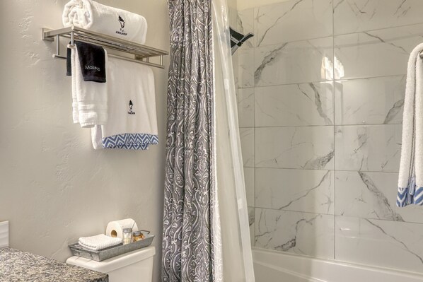 A bathroom with a marble shower wall, a towel rack with multiple towels, a sink with toiletries, and a patterned shower curtain.