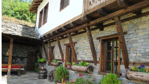 Ein traditionelles Steinhaus mit Holzbalken und Fenstern. Übertöpfe mit Pflanzen und ein Holzwagen mit Pflanzen sind ebenfalls zu sehen.