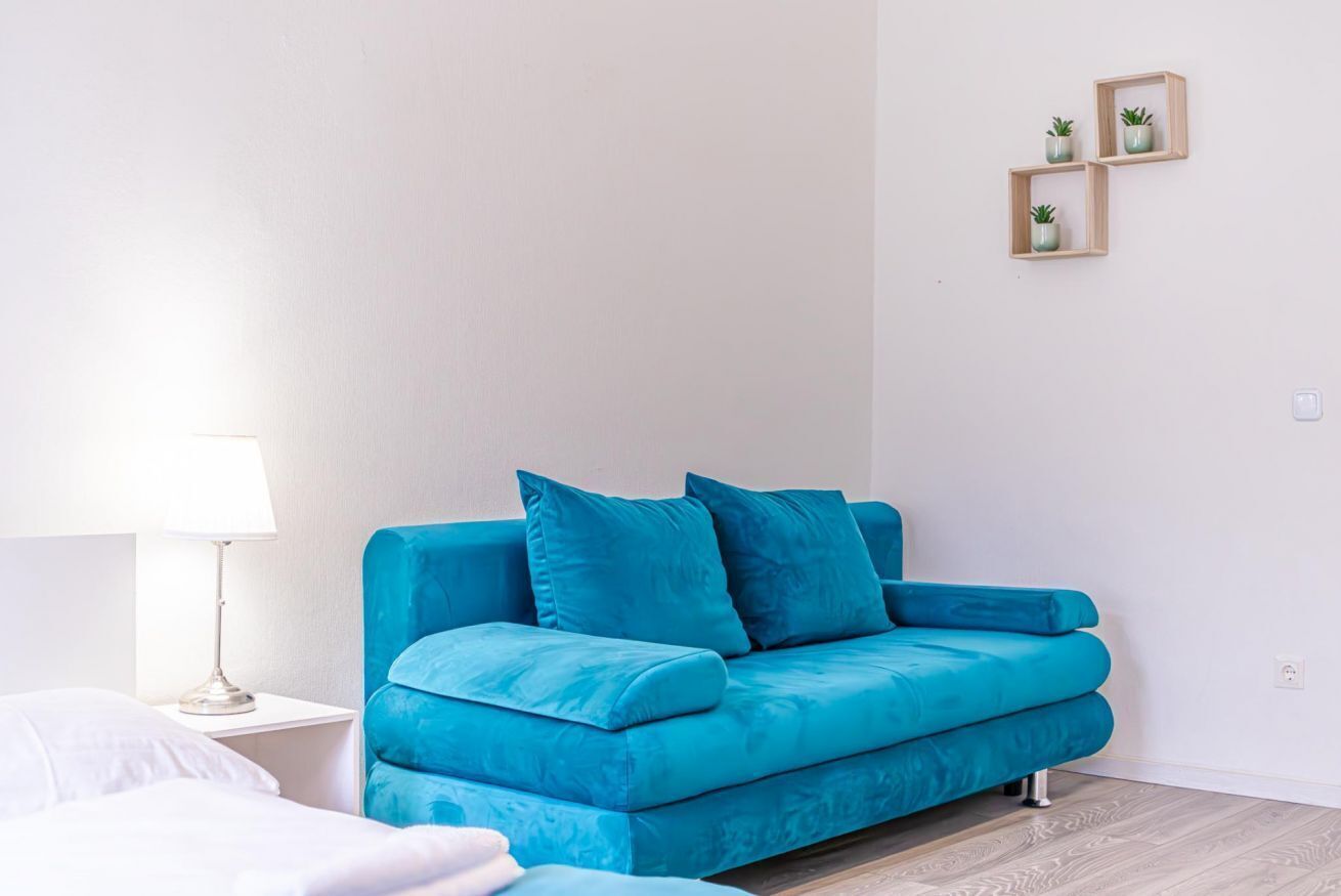 Une chambre moderne avec un canapé de couleur turquoise, une table de chevet blanche, une lampe et une petite étagère en bois ornée de plantes.
