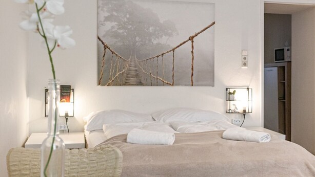 A neatly made bed with white linens, a wall-mounted picture of a bridge, bedside lamps, and a small bedside table with a vase of flowers.
