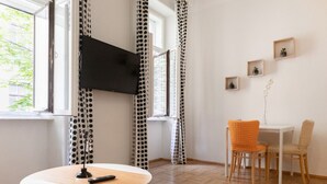 A room with a TV, a round table, two chairs, a window with curtains, and a wall with framed pictures.