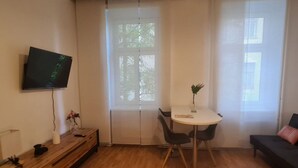 A modern living room with a TV mounted on the wall, a dining table with chairs, a sofa, and a window with blinds. - Cozy City Center Retreat 2BR Apt. Homelike Comfort (Vienna)