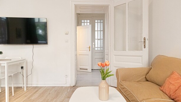 Una sala de estar con un televisor de pantalla plana, una mesa blanca con un florero, un sofá color beige y una puerta que da a otra habitación.