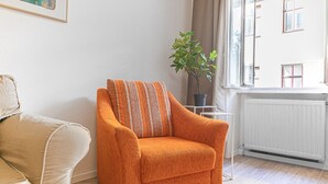 Una habitación con un sofá color beige, un sillón naranja con cojines a rayas, una planta en maceta y una ventana con vista a edificios.