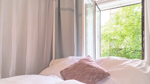 Una cama con ropa de cama blanca y una almohada rosa, una ventana con cortinas translúcidas y vistas a follaje verde afuera.